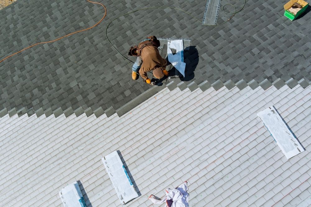 Roofer on a roof, installing gray shingles. Overhead view, partially finished with white shingles below.