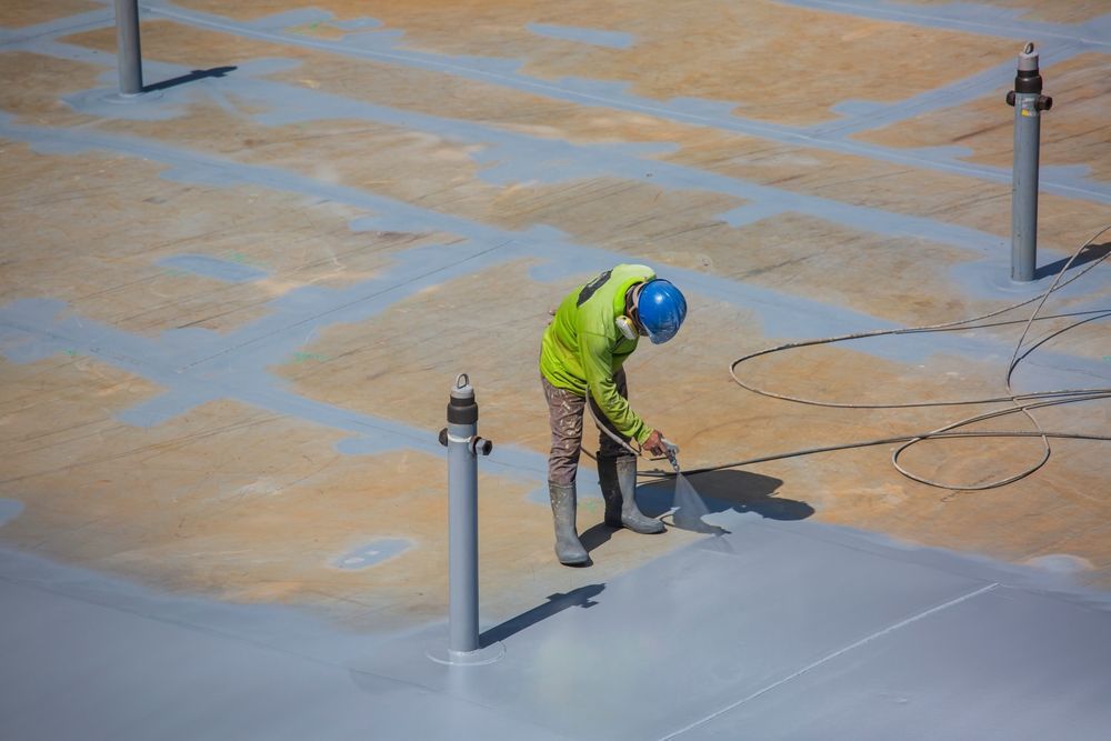Construction worker in safety gear spraying a grey coating on a large surface outdoors.