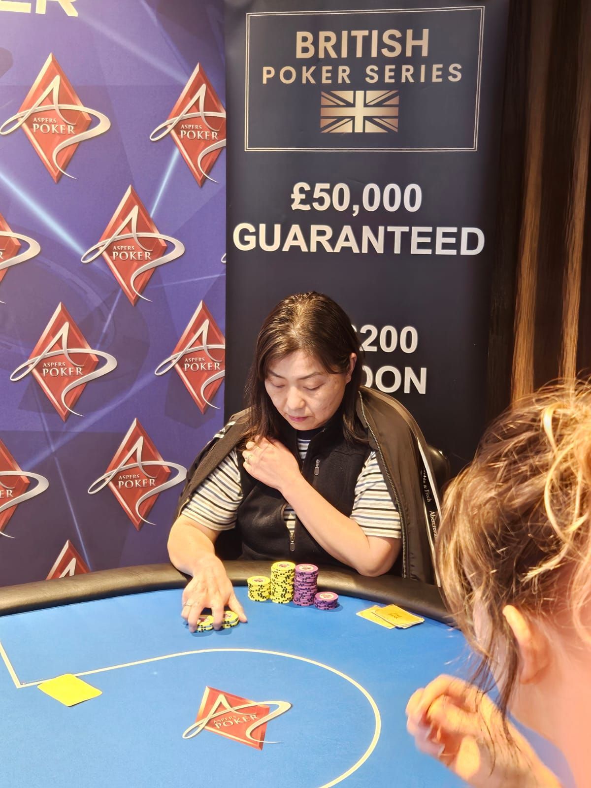 Woman playing poker at a table, looking down at cards and chips, at a British Poker Series event.