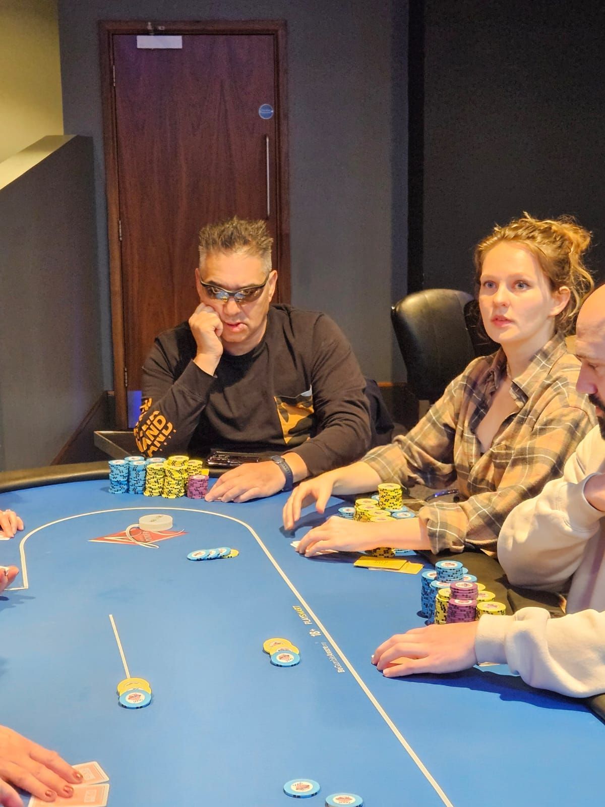 People playing poker at a table. Woman in plaid shirt, man in sunglasses; chips on the felt.