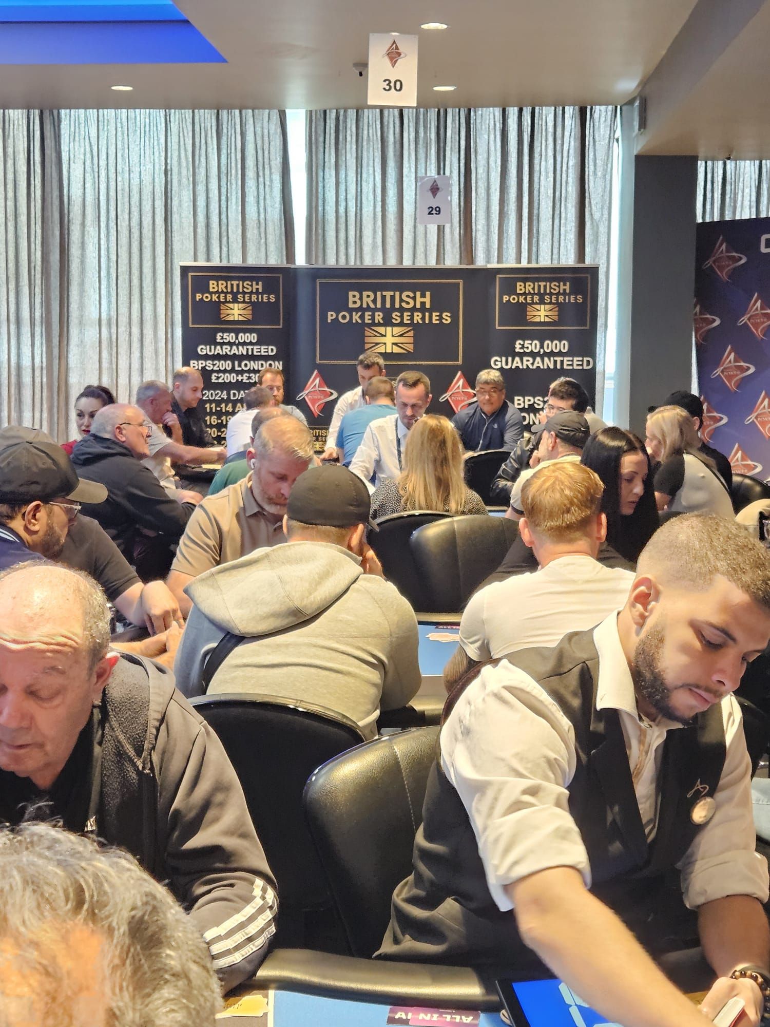 Poker tournament in progress; players seated around tables, focused, with British Poker Open banners visible.