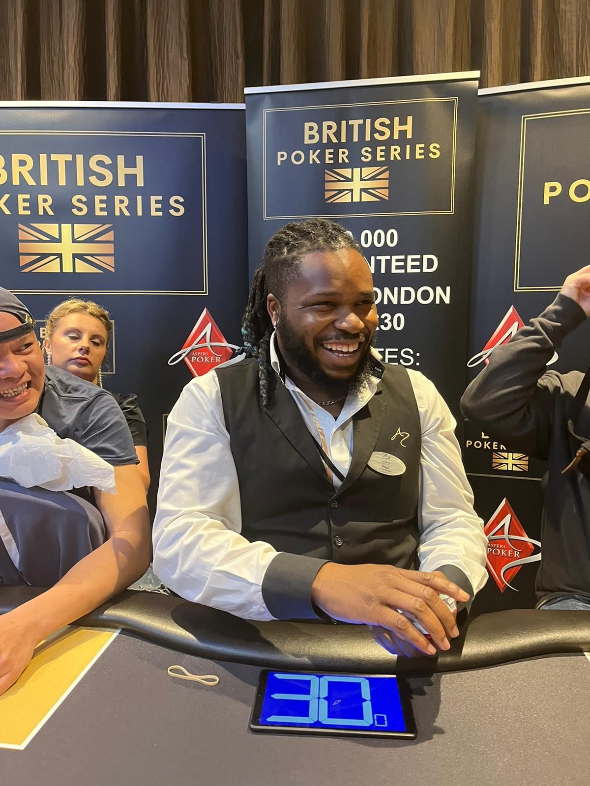 Man in vest smiles at poker table, flanked by others. 