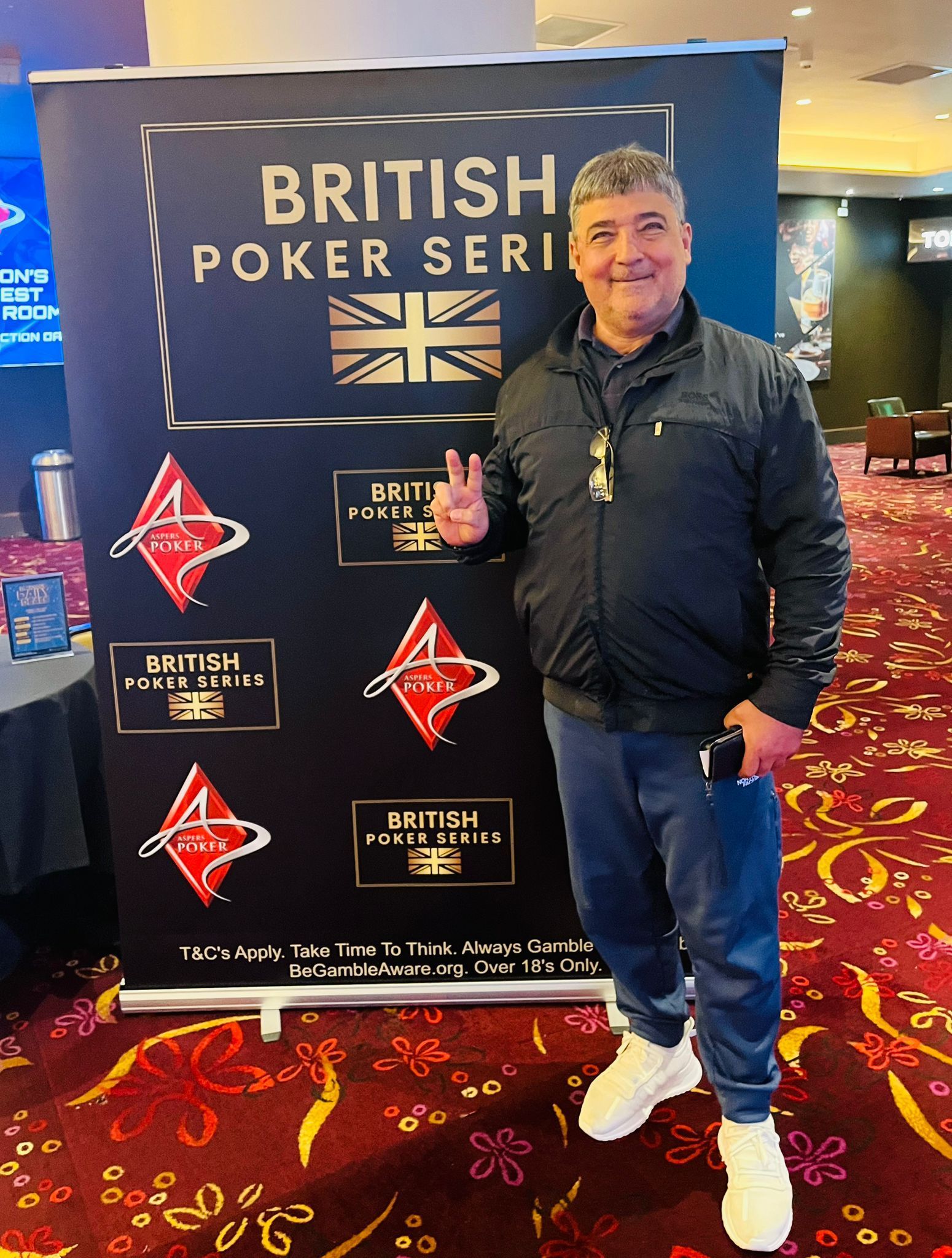 Man posing in front of British Poker Series banner, wearing jacket and jeans, flashing a peace sign.