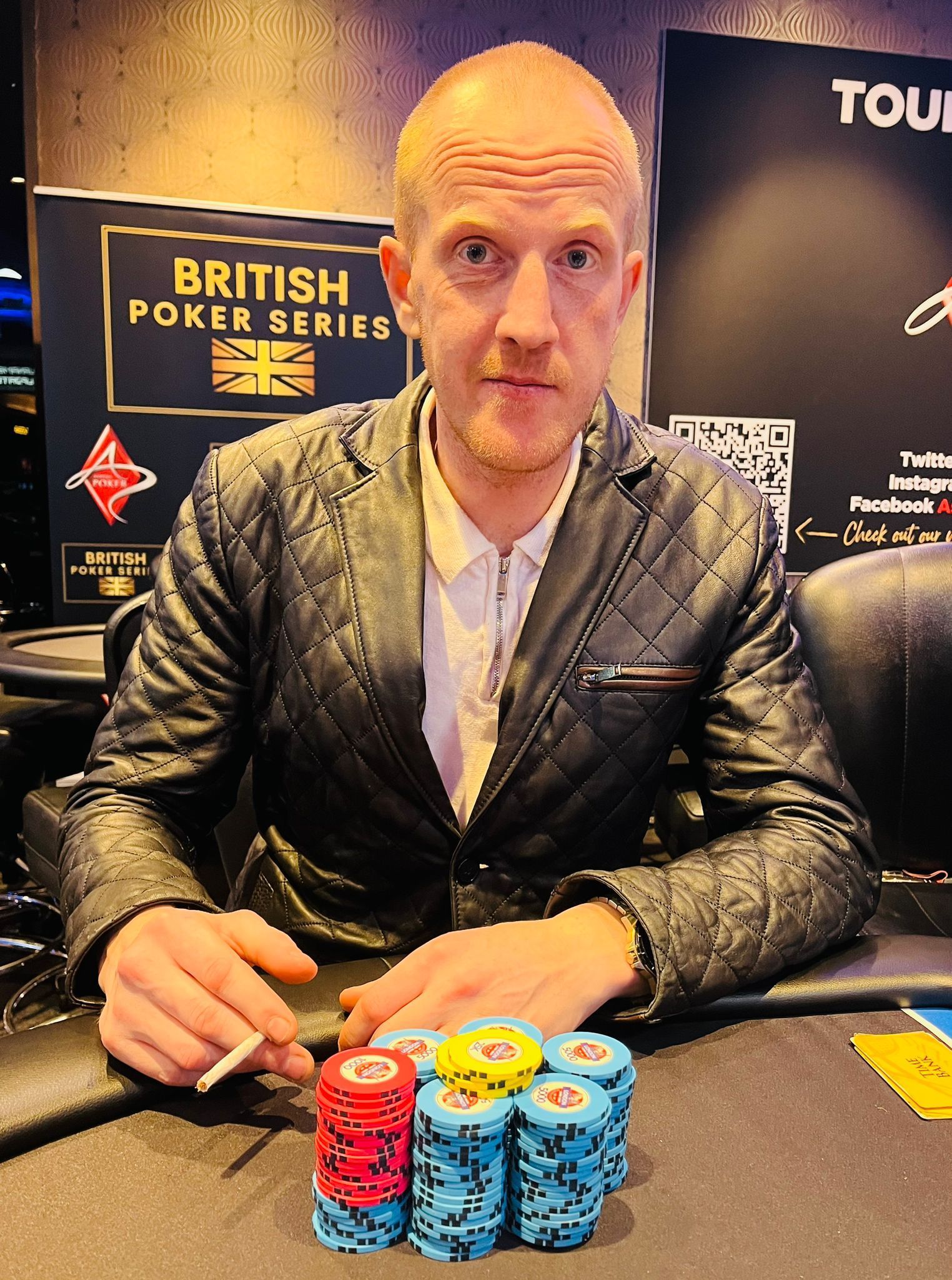 Man at poker table with chip stacks, smoking, wearing jacket. British Poker Series banner visible.