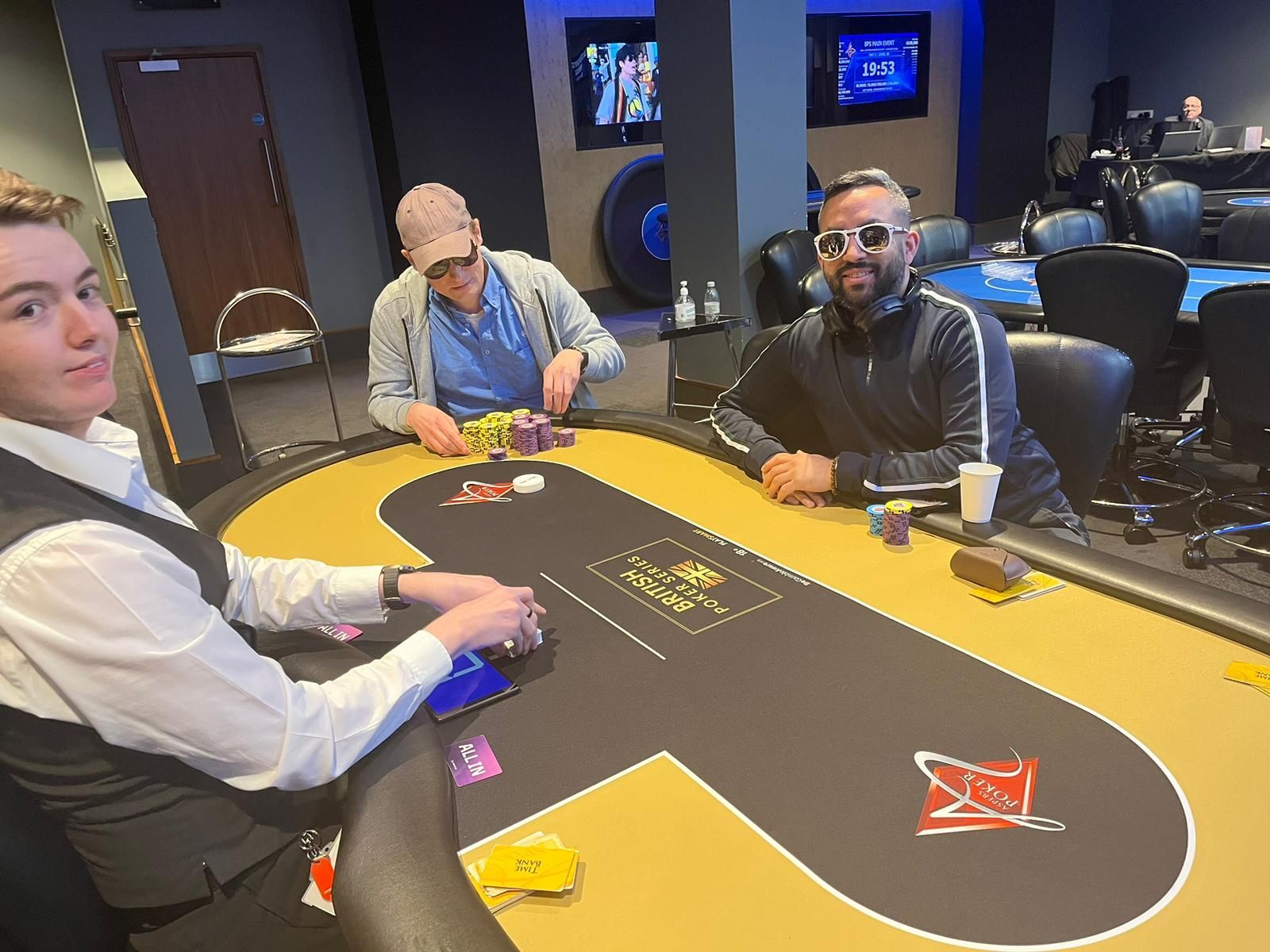 Three people playing poker at a table in a casino. Man in sunglasses smiles, others concentrate on cards and chips.