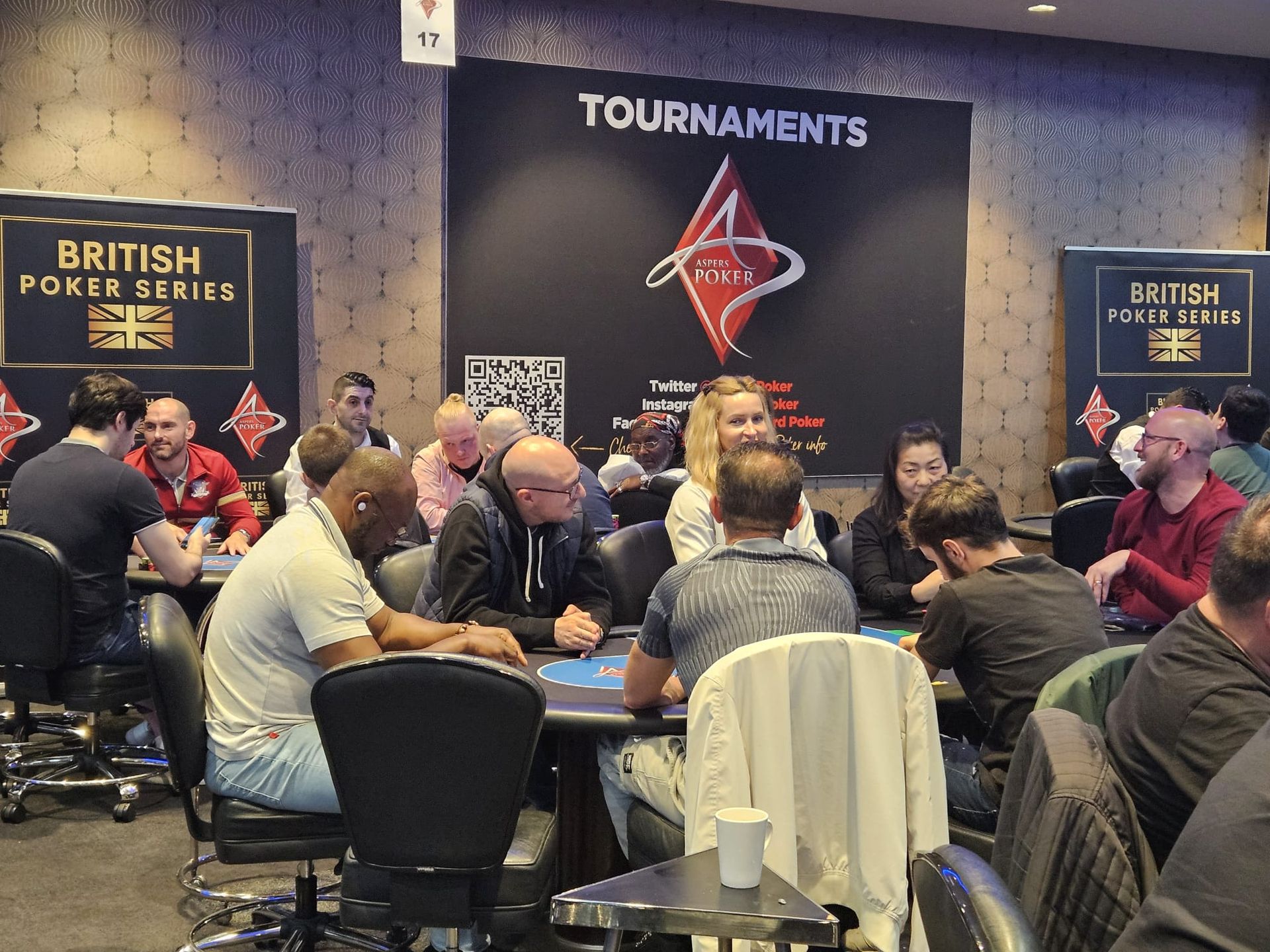 Poker tournament in progress, players seated at tables, casino setting with British Poker Series branding.