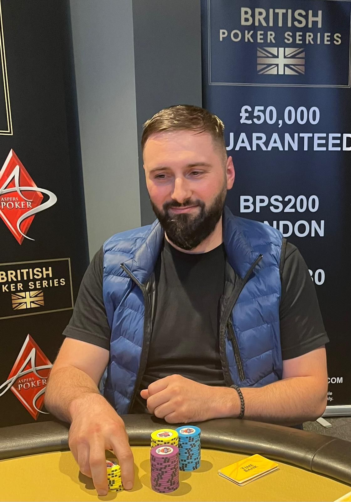 Man at poker table, smiling, wearing blue vest, cards in front of him. British Poker Series backdrop.