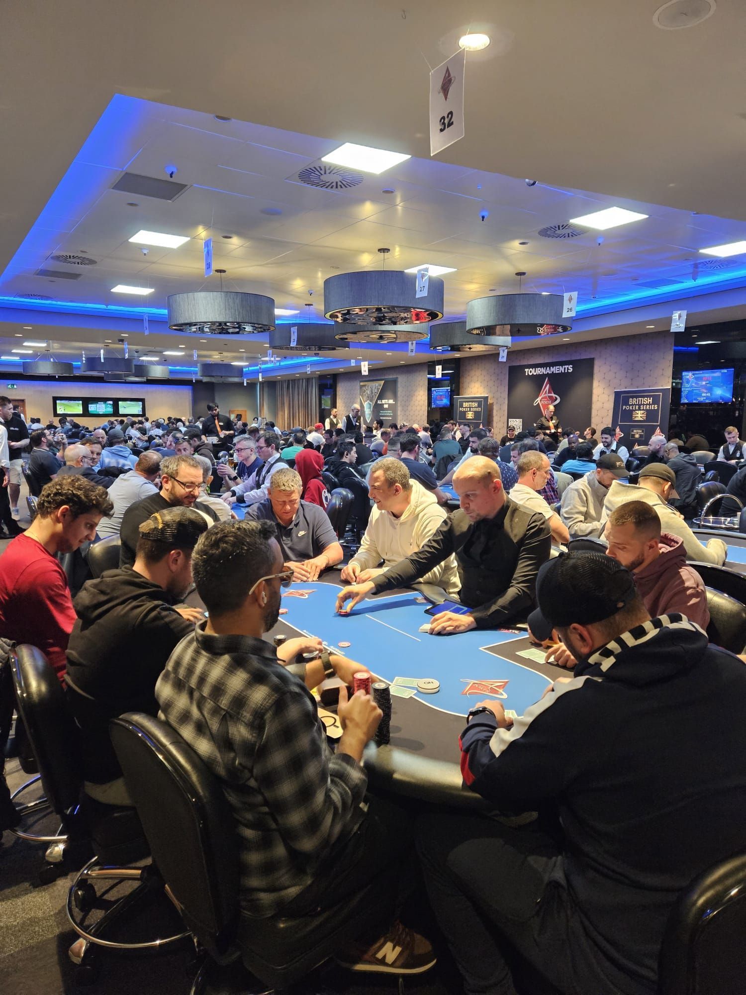 Poker players seated at tables in a casino, bright overhead lighting.