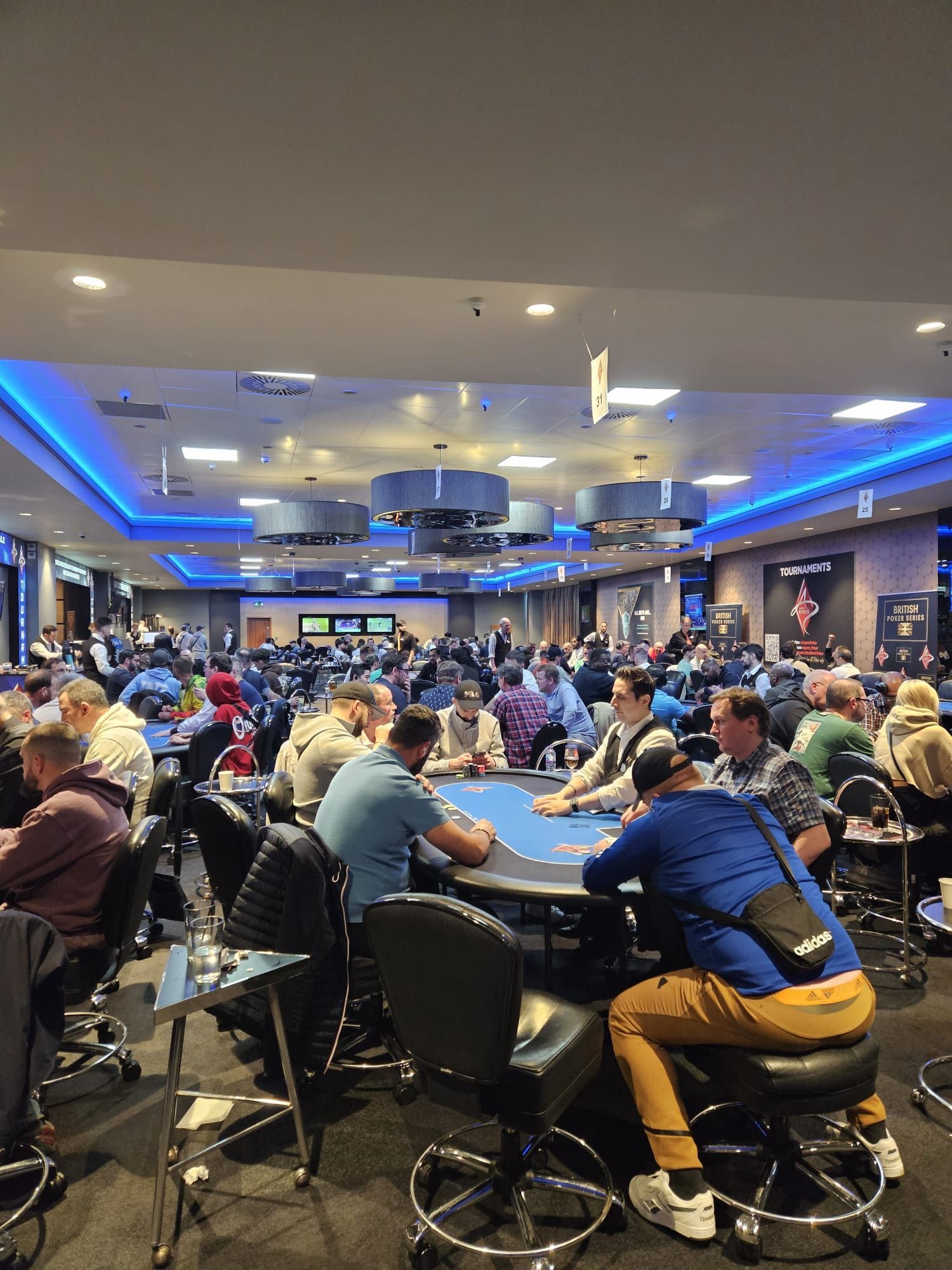 Poker room with many players seated at tables, lit by blue and overhead lights.