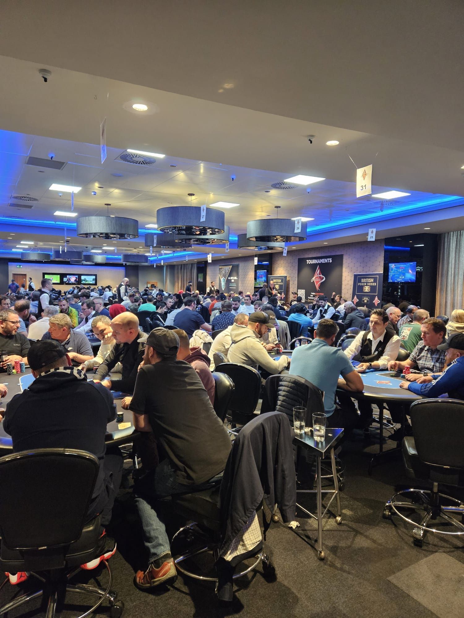 Large poker room filled with players seated at tables, lit with blue and white lights.