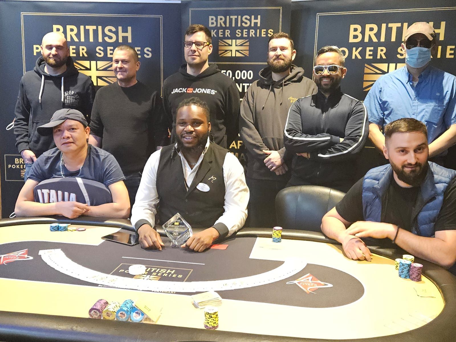 Group of poker players pose at a table, some with arms crossed, trophy in center. 
