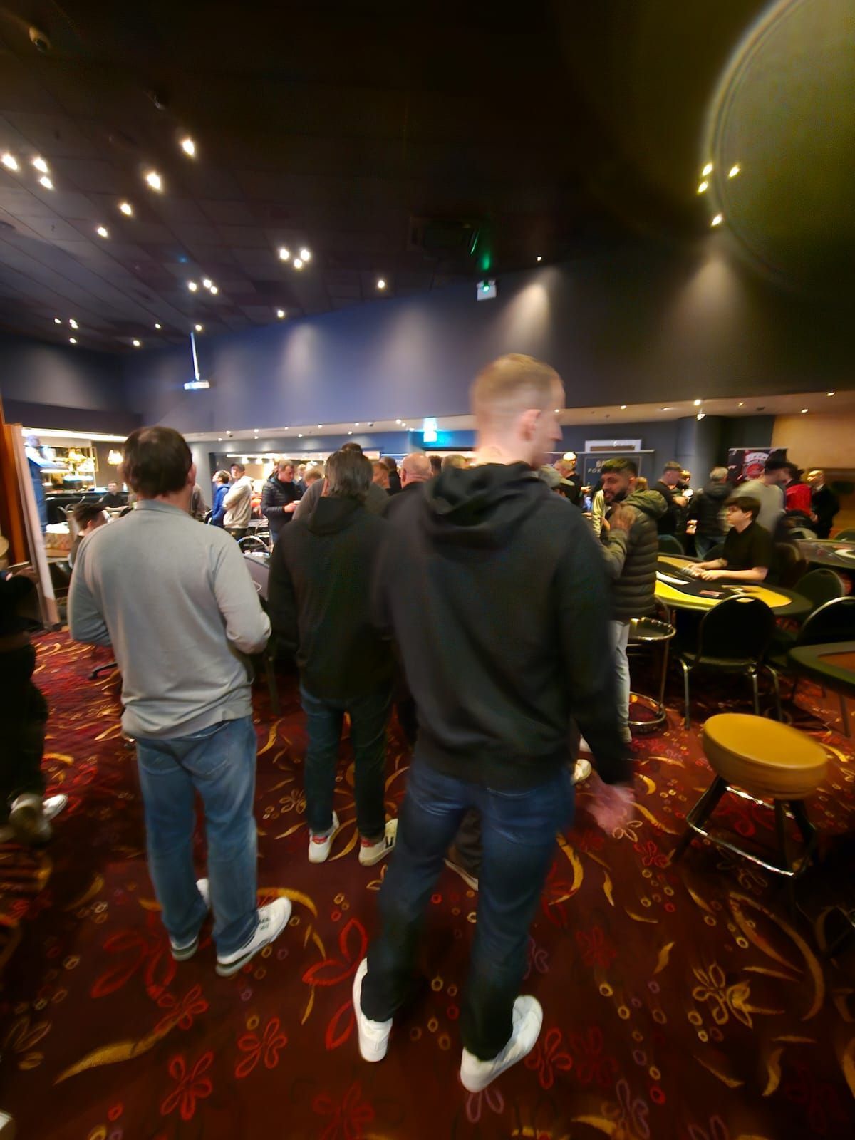Men in casual clothes in a crowded room with tables. Red patterned carpet and overhead lights.