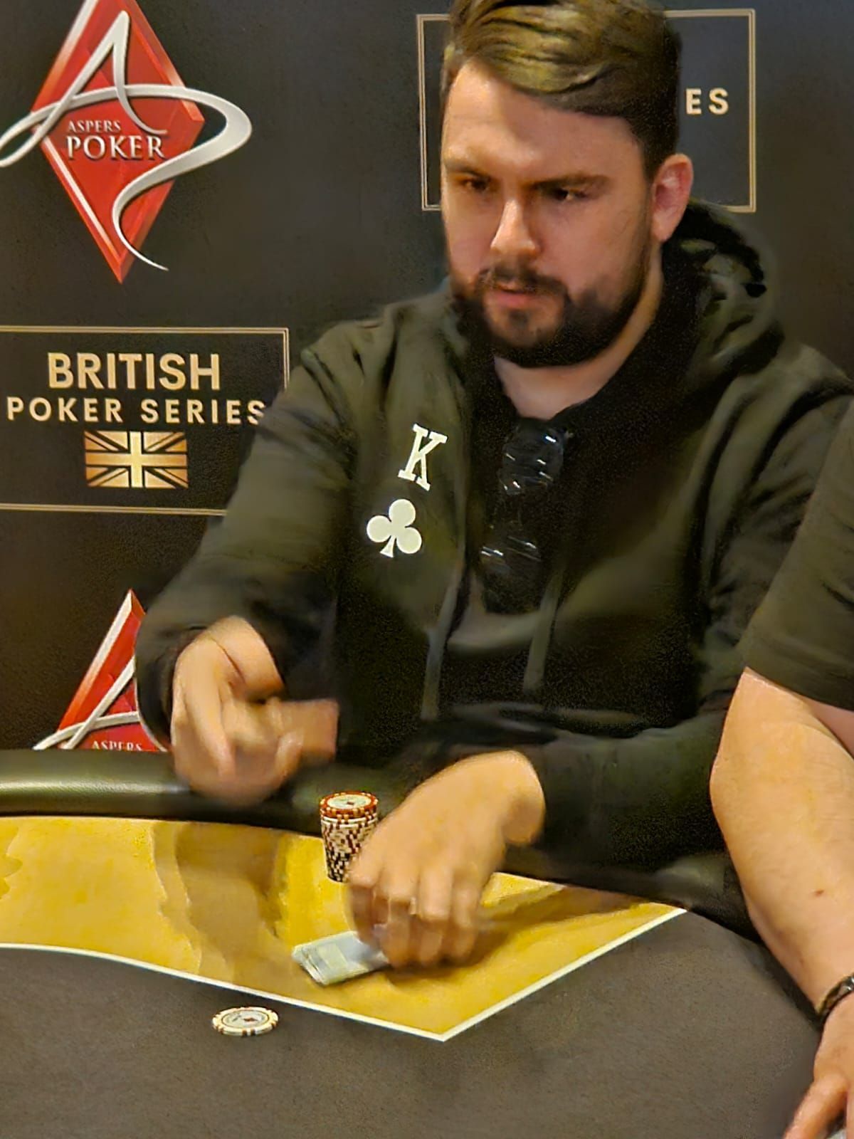 Man at poker table with cards, wearing a black hoodie with a King of Clubs patch. British Poker Series logo in the background.