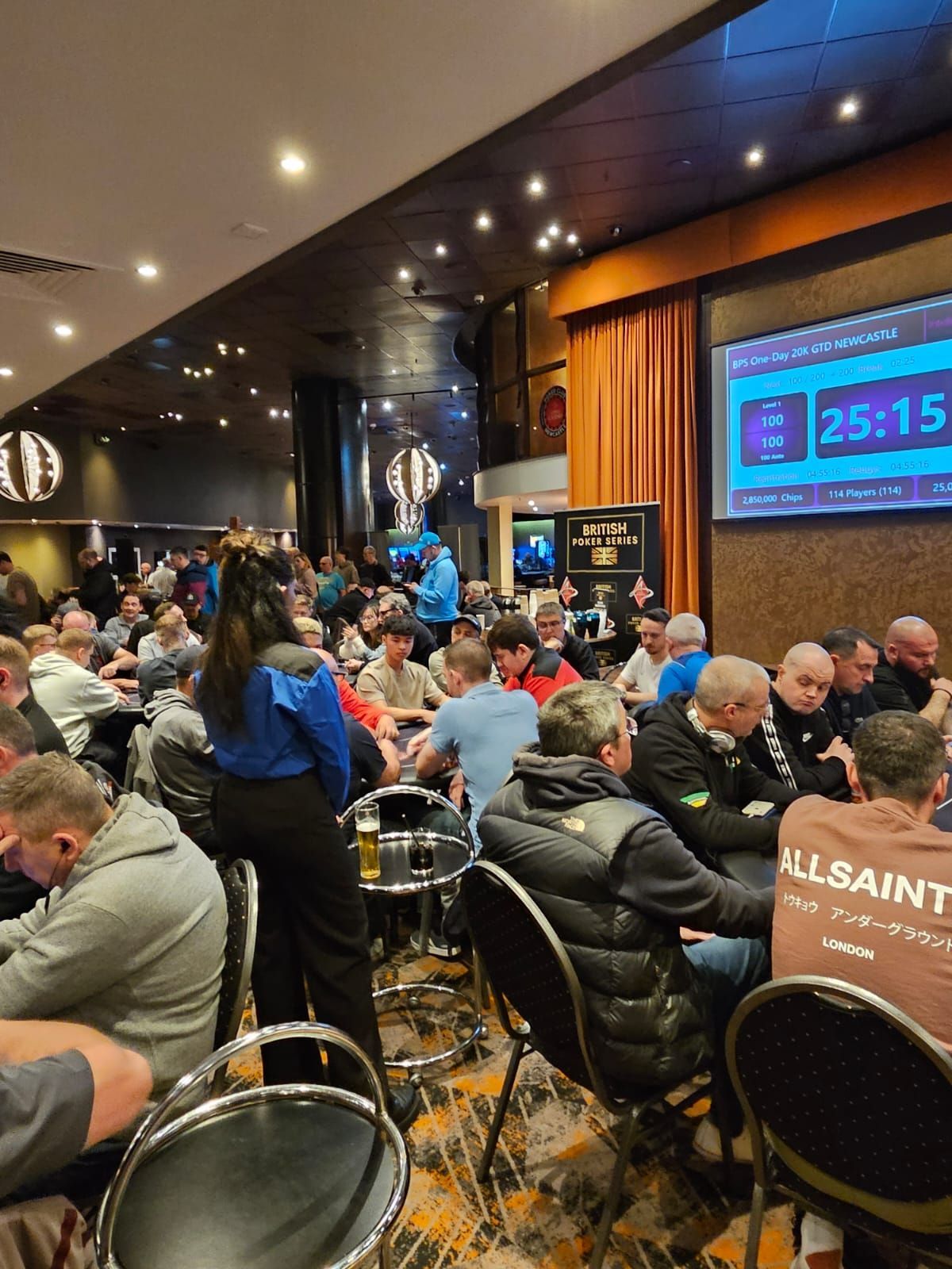 Poker tournament in a crowded room. People seated at tables, a timer visible.