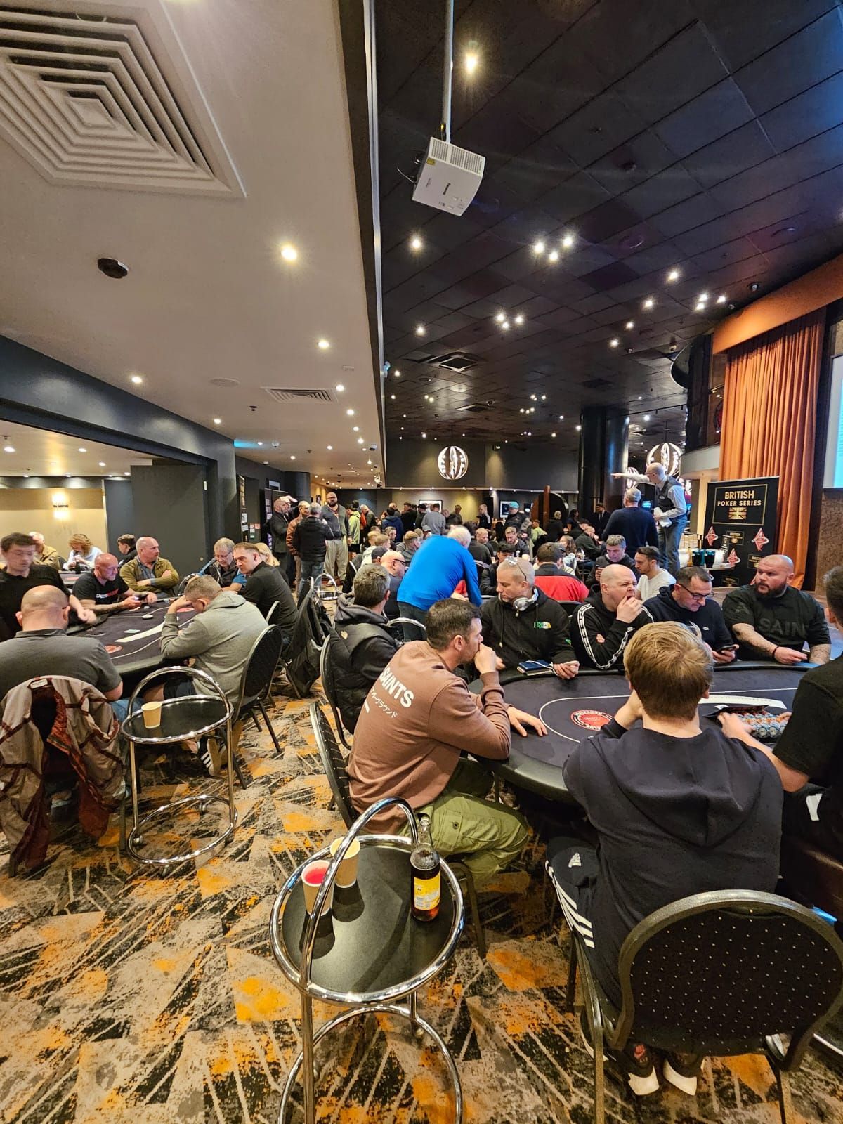 Poker room with numerous people at tables, lit by overhead lights. Dark decor with a busy atmosphere.
