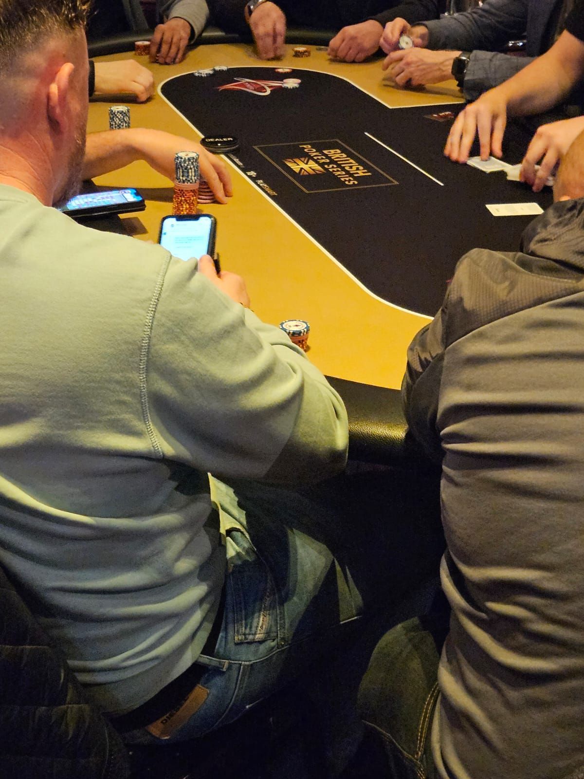 Poker game in progress at a table. People seated around the table, one using phone, cards visible.