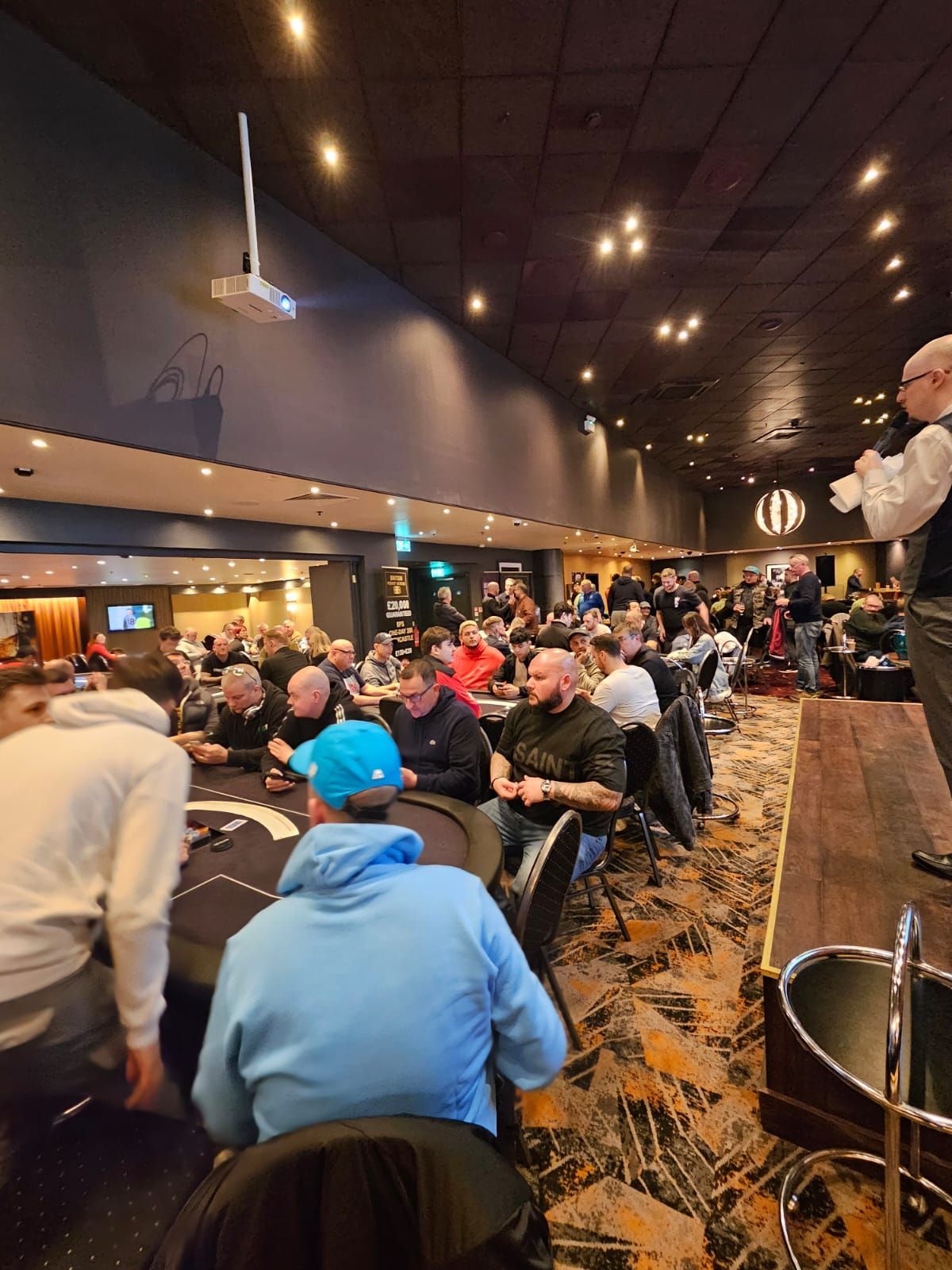 Poker tournament in progress, many people seated at tables in a large room, announcer at front.