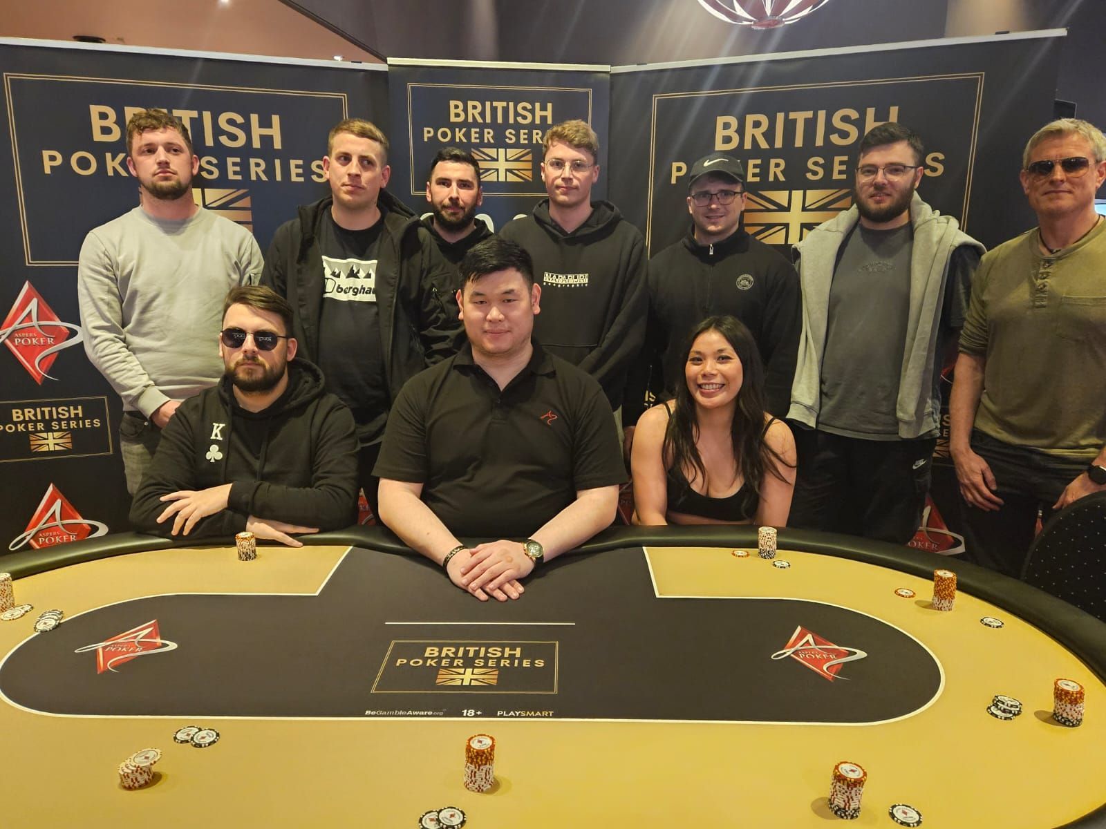 Poker players at a table, posing for a group photo. British Poker Series banner visible.