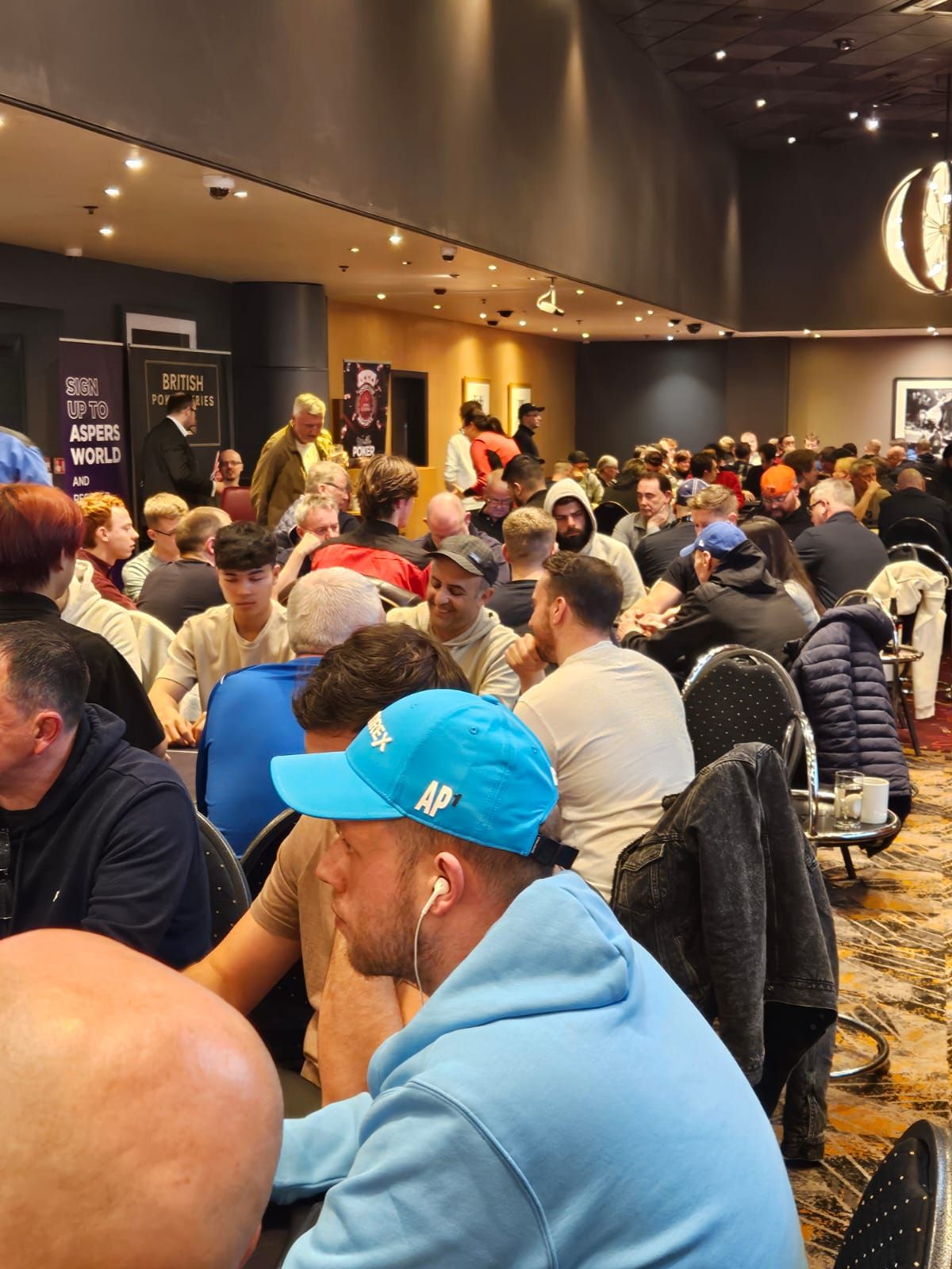 A crowded poker room with many people seated at tables, some wearing hats.