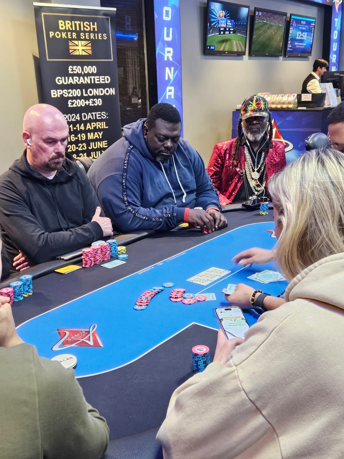 Poker players at a table, cards dealt. A man in a red jacket, others focused, chips visible. Tournament banner in background.