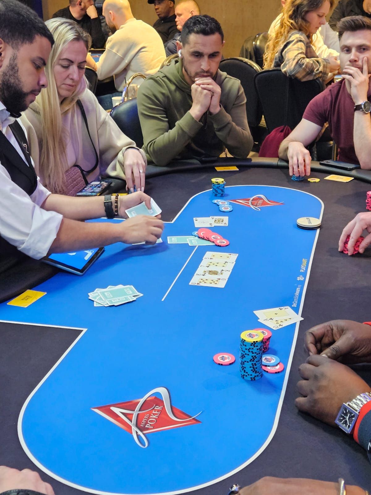 Poker game in progress. Players at a blue felt table, cards visible, chips stacked. Men in focus, observing.