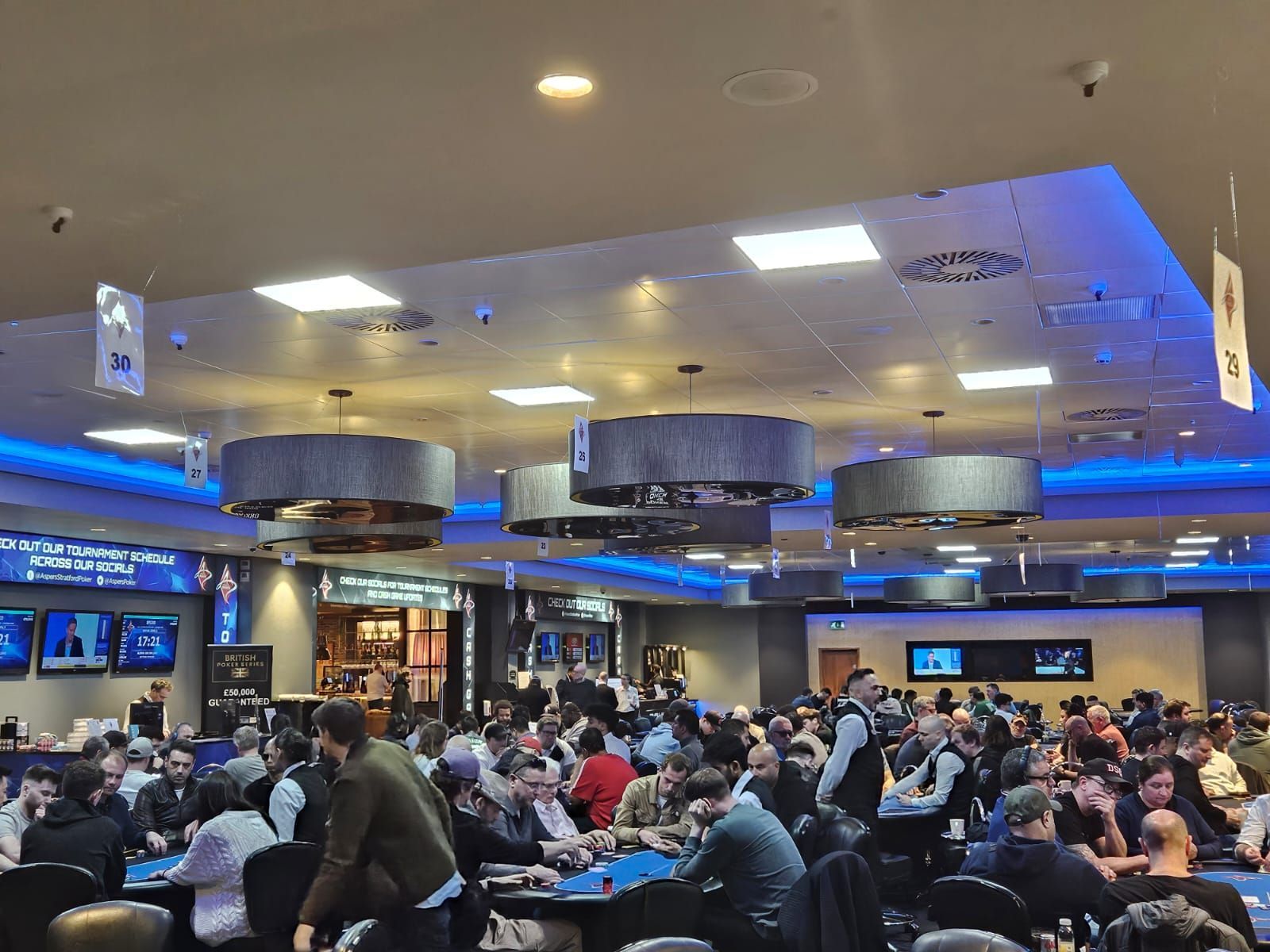 Crowded casino interior with poker tables, players, and staff; overhead lighting.