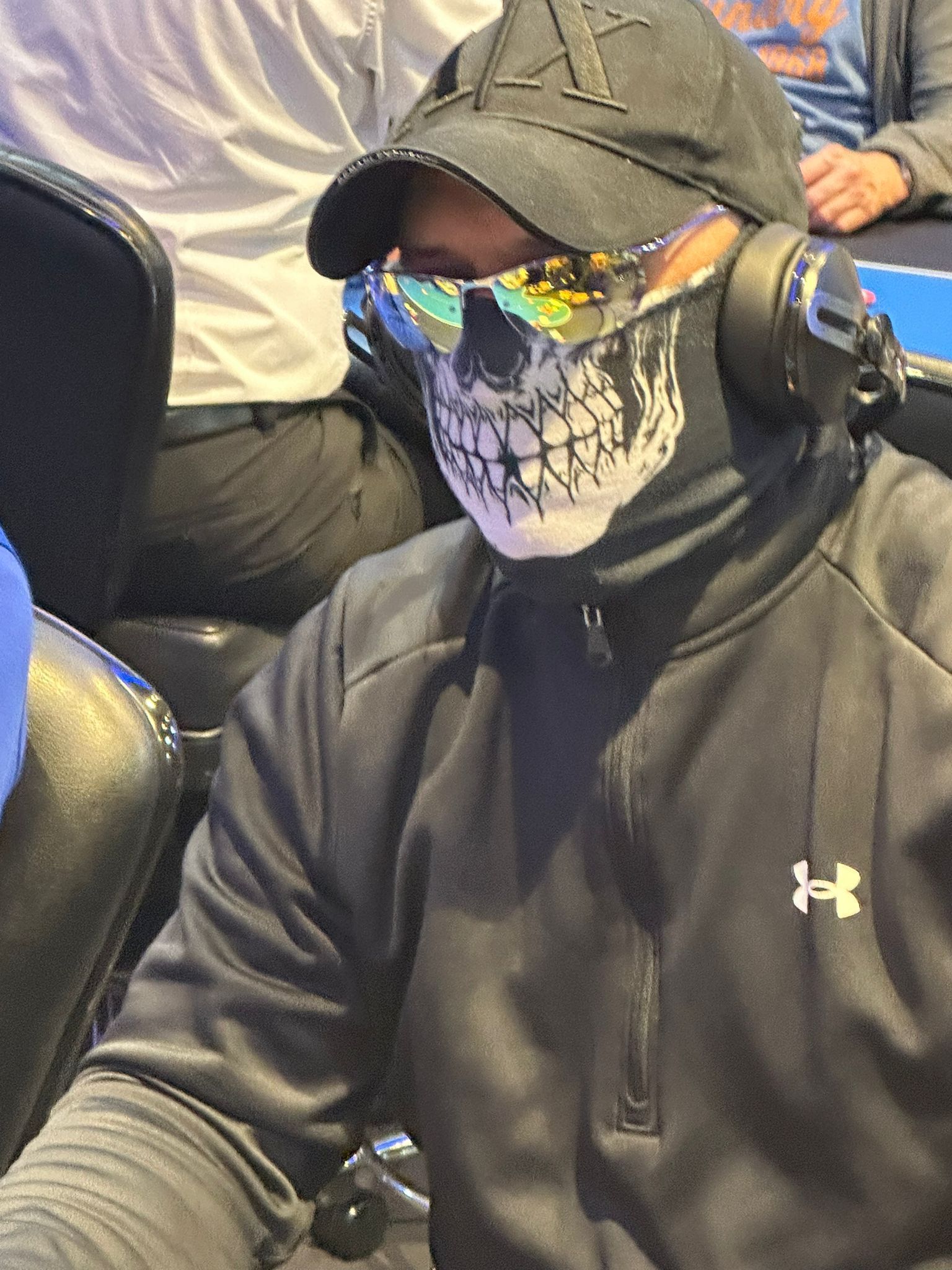 Person wearing a skull mask, sunglasses, and a baseball cap, sitting indoors, likely at an event.