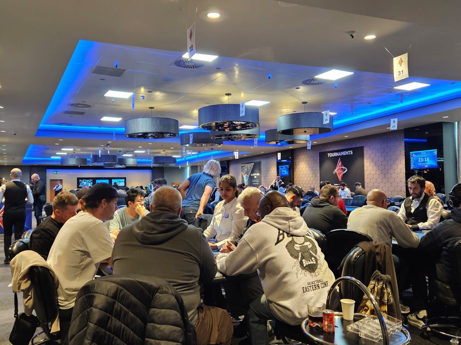Poker room with people seated at tables, playing cards under bright lights.