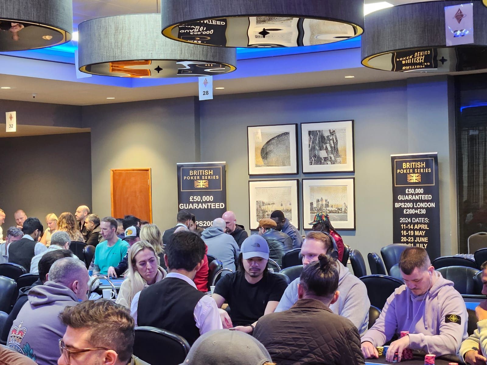 Poker room with people seated at tables, playing cards. Dark lighting, posters on walls.