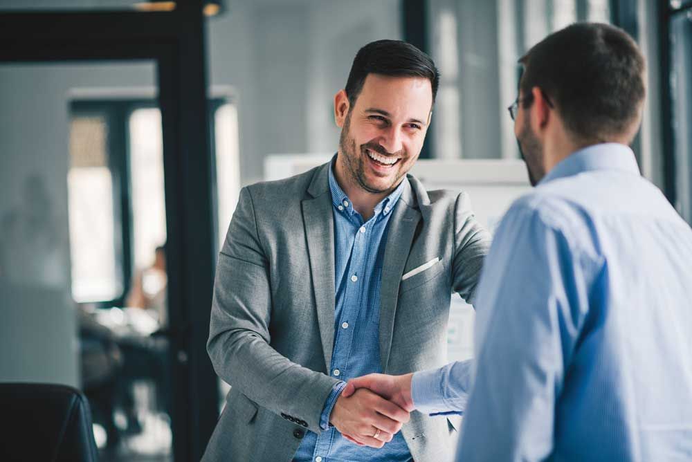 Two men are shaking hands in an office and smiling.