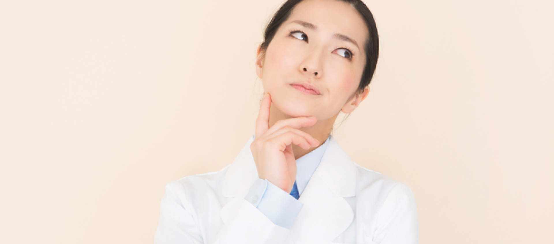 A woman in a lab coat is looking up with her hand on her chin.