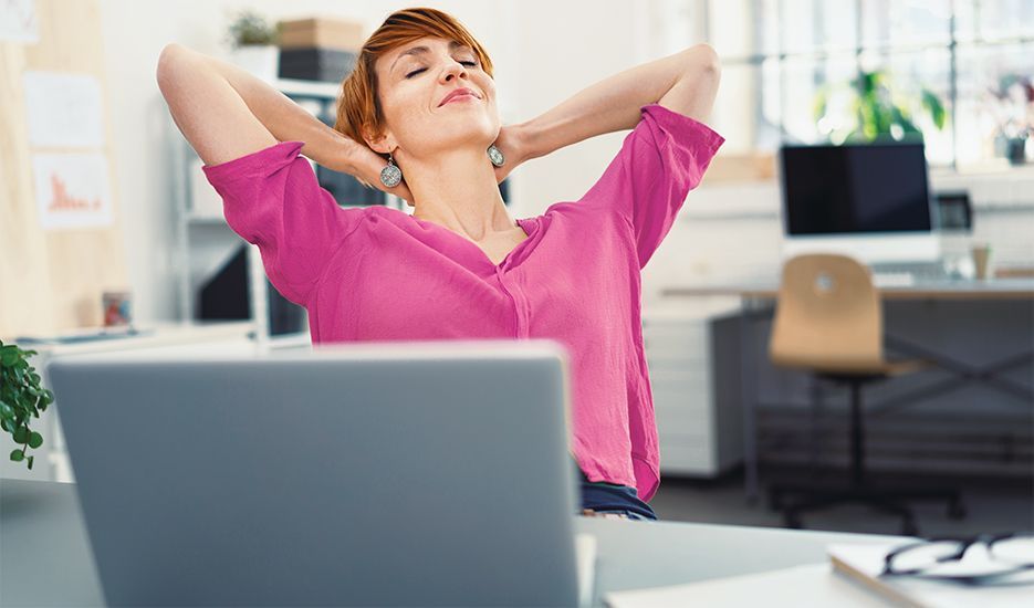 A woman is sitting at a desk with her hands behind her head in front of a laptop computer.