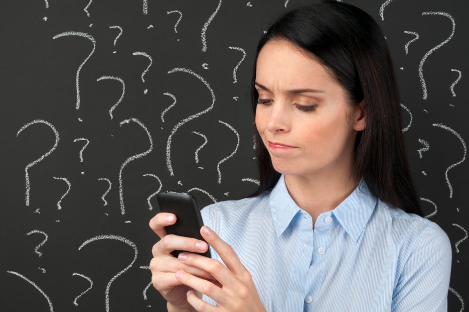 A woman is holding a cell phone in front of a blackboard with question marks drawn on it.