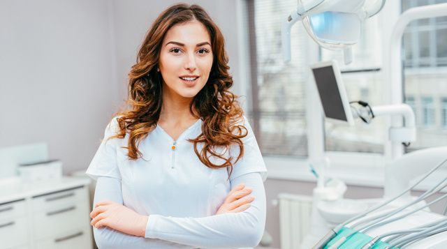 A female dentist is standing in a dental office with her arms crossed.