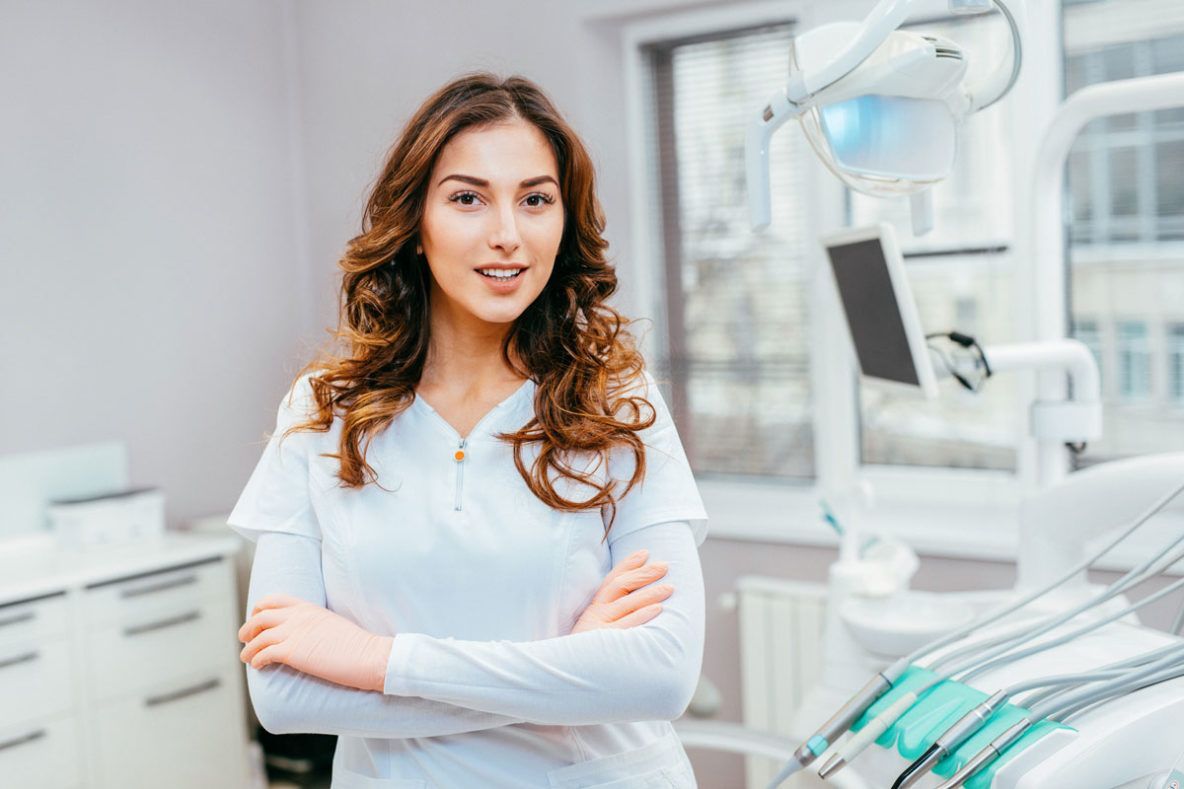 A female dentist is standing in a dental office with her arms crossed.