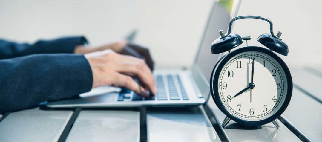 A person is typing on a laptop next to an alarm clock.