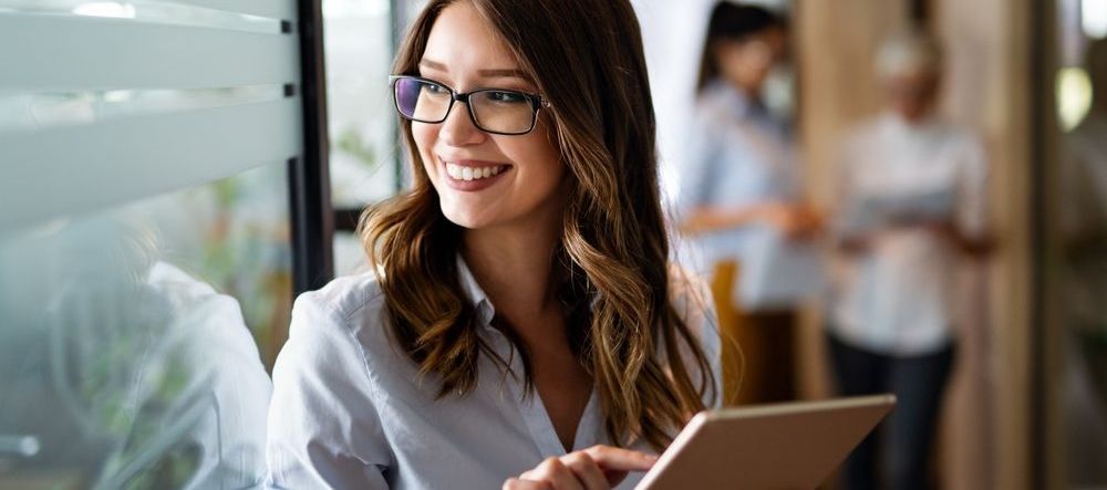 A woman wearing glasses is holding a tablet and smiling.