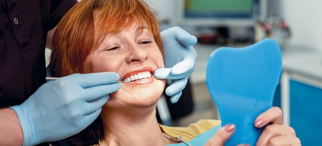 A woman is sitting in a dental chair looking at her teeth in a mirror.