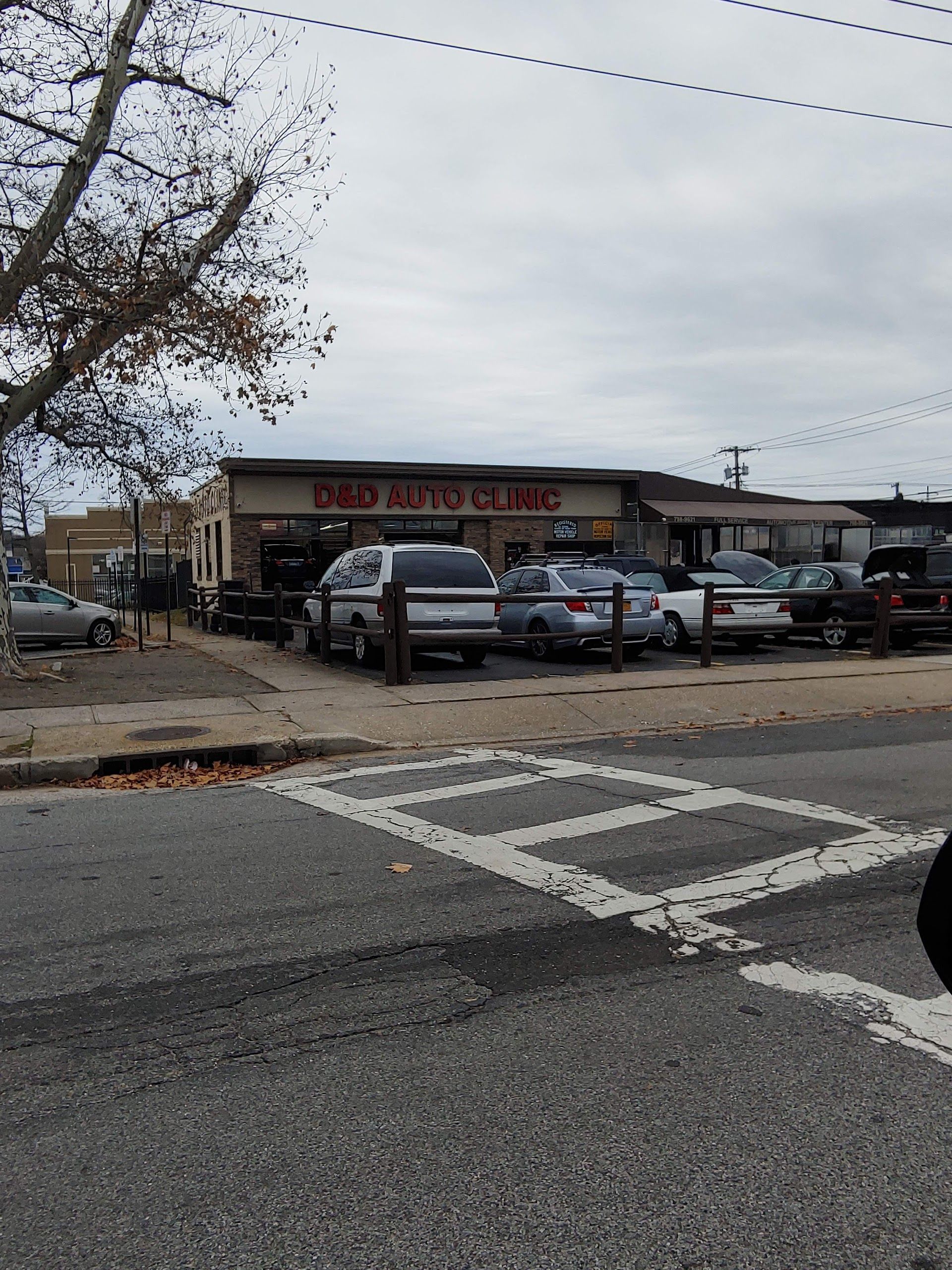 D&D Auto Clinic building with cars parked in front, viewed from a crosswalk on a cloudy day. | D & D Auto Clinic