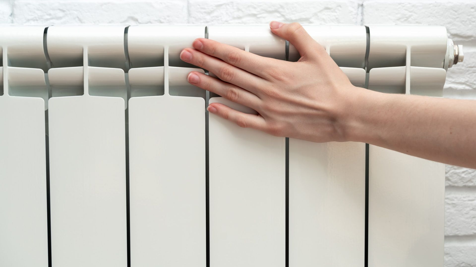Eine Hand ruht auf einem weißen Metallheizkörper, der an einer weißen Ziegelwand befestigt ist.