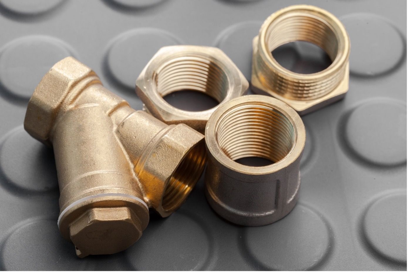 Ein Y-förmiges Siebventil aus Messing und mehrere Gewindefittings sind auf einer grauen, strukturierten Gummimatte angeordnet.