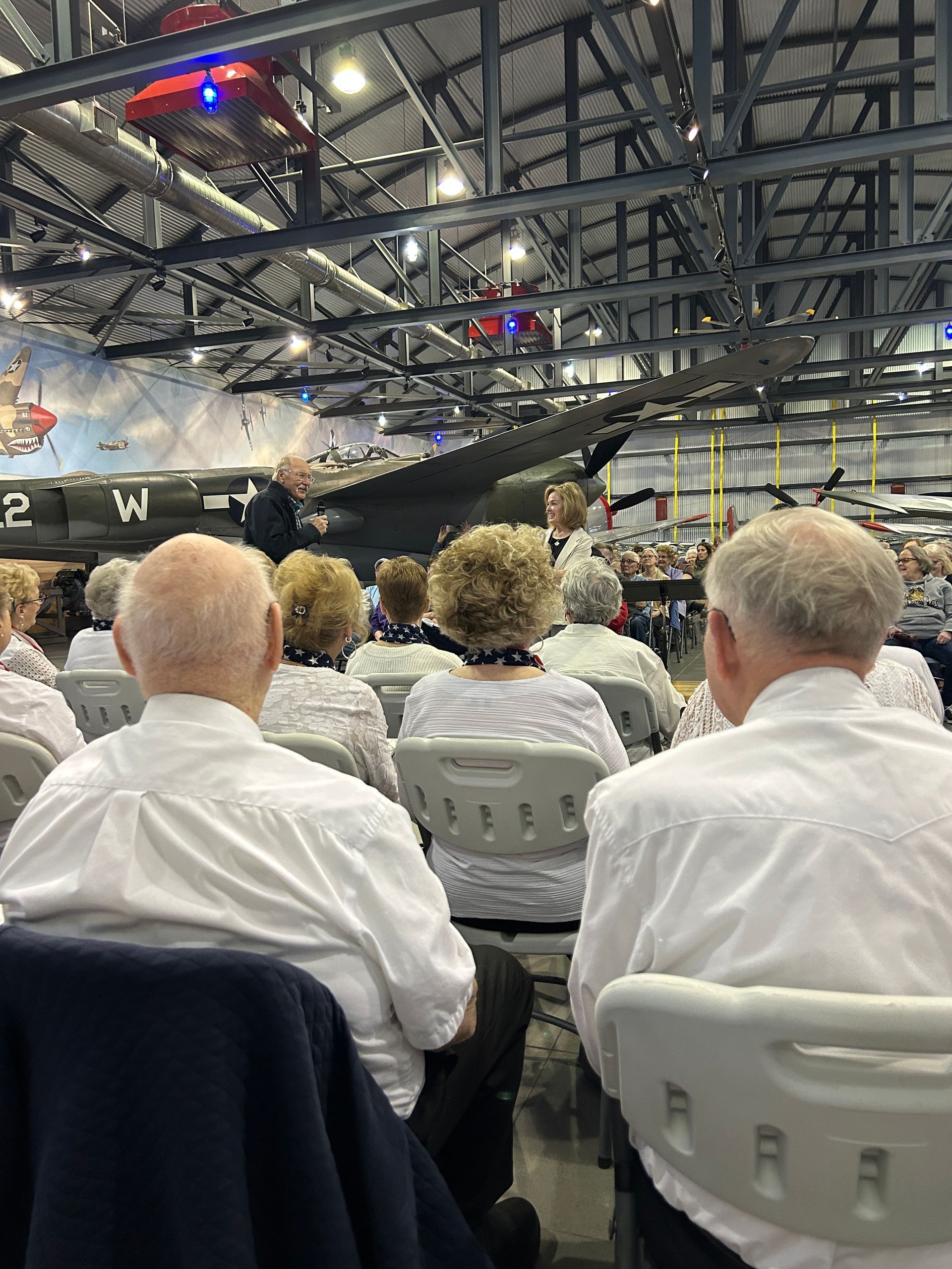 People seated, listening to a presentation in an aircraft hangar. Aircraft visible in background.