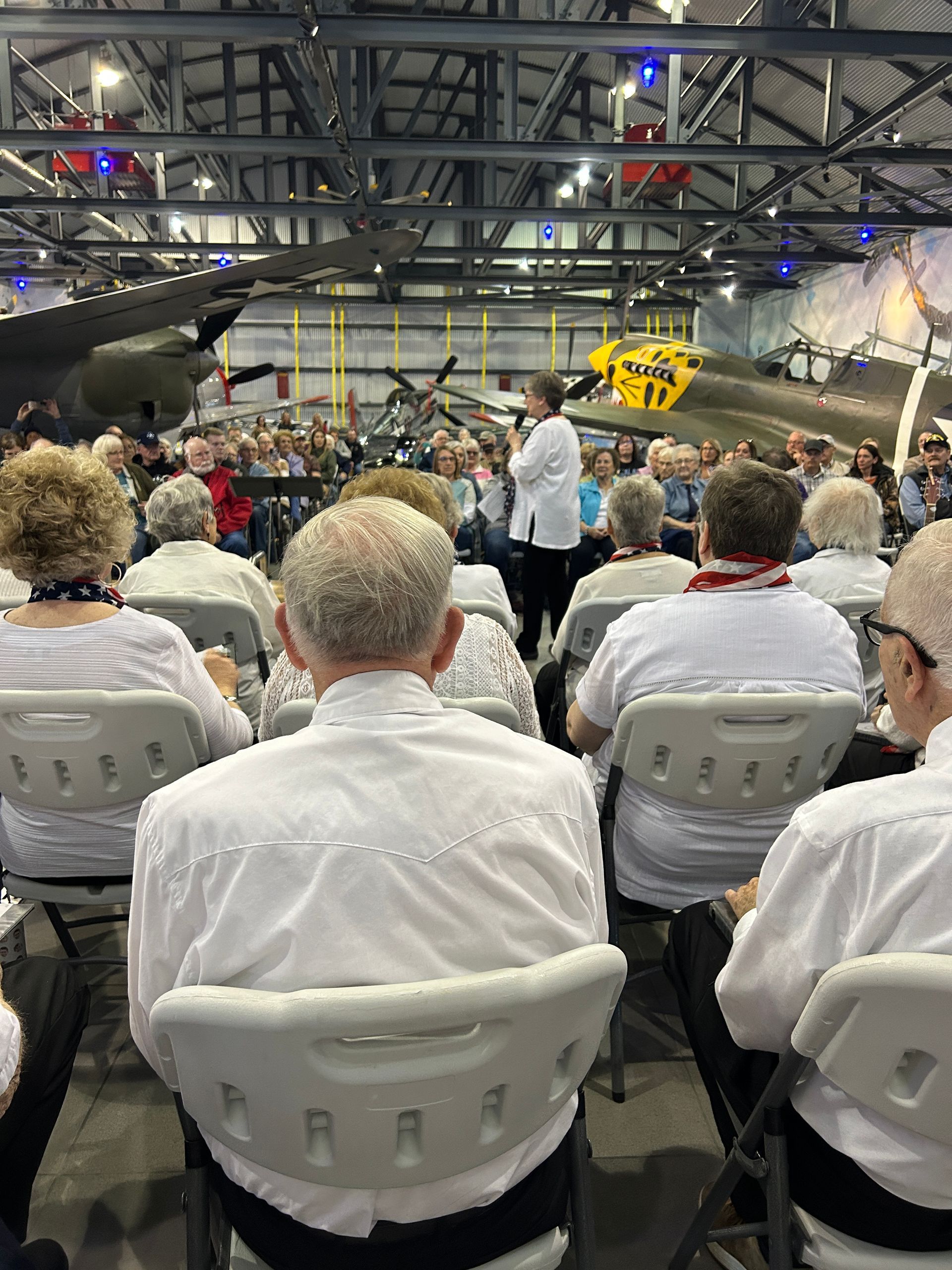 Audience seated, listening to a speaker in an airplane hangar. Airplanes are visible in the background.
