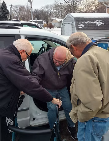 Three men assisting another man to exit a white van, snowy outdoor setting.