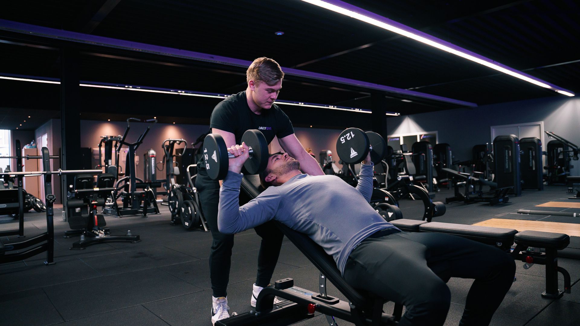 Een man helpt een andere man met het tillen van halters in een sportschool.