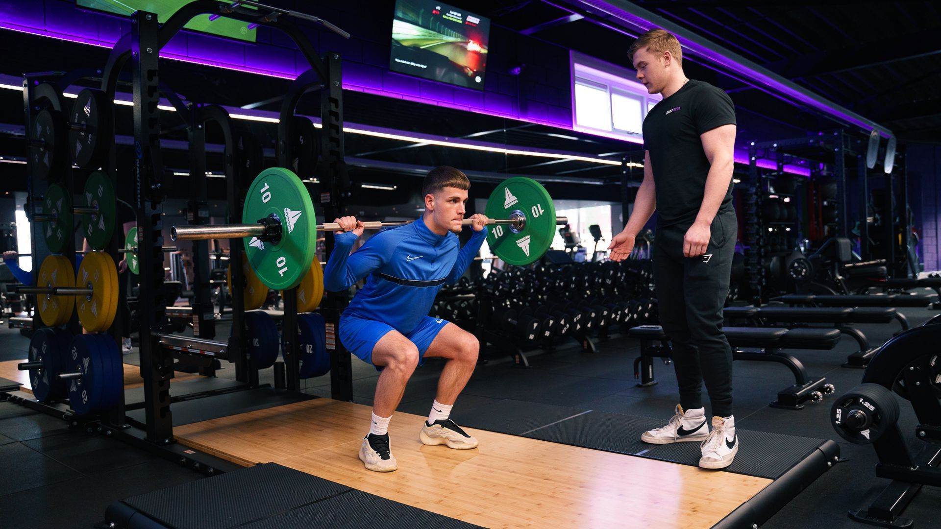 Een man hurkt met een halter in een sportschool, terwijl een andere man toekijkt.