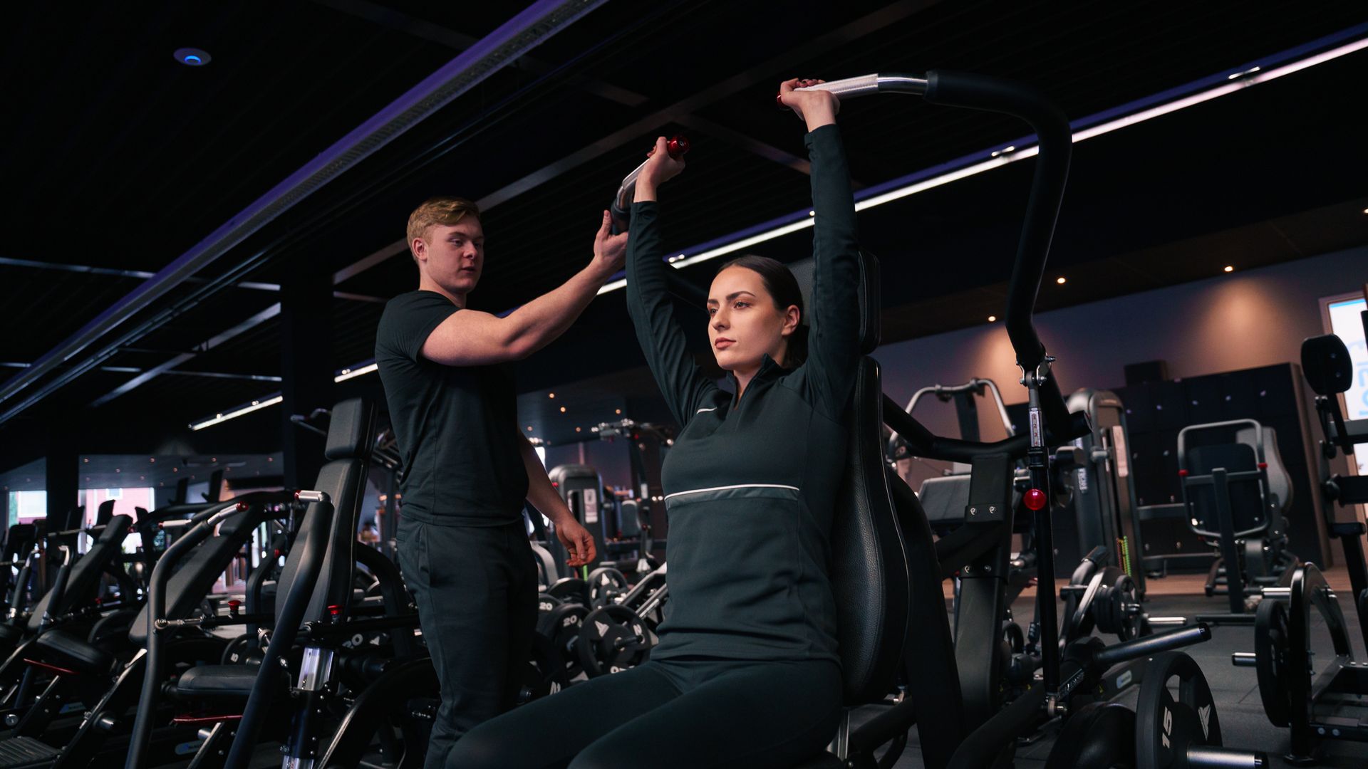Vrouw die onder begeleiding van een trainer in een sportschool een schouderpersapparaat in zittende positie gebruikt.
