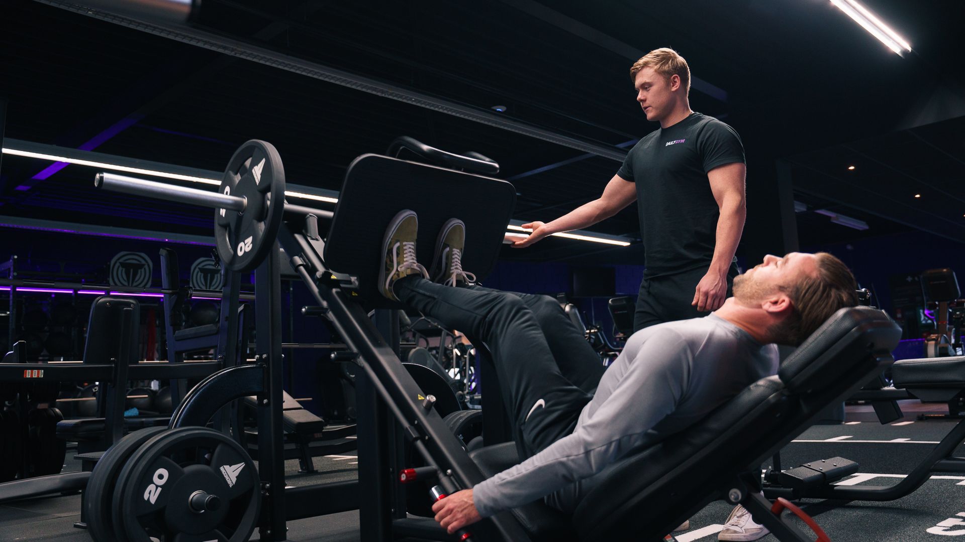 Een man gebruikt een legpress-apparaat in de sportschool. Een trainer kijkt toe en corrigeert zijn houding.