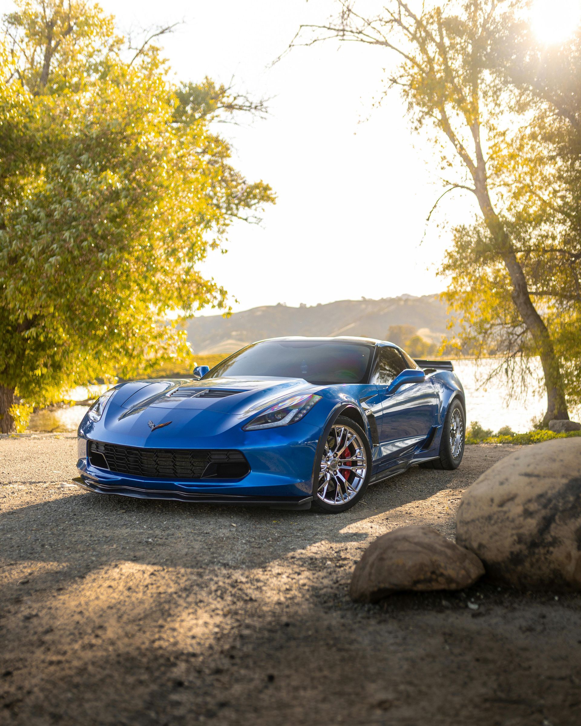 Blue Corvette sports car parked on a dirt road near a lake, surrounded by trees.