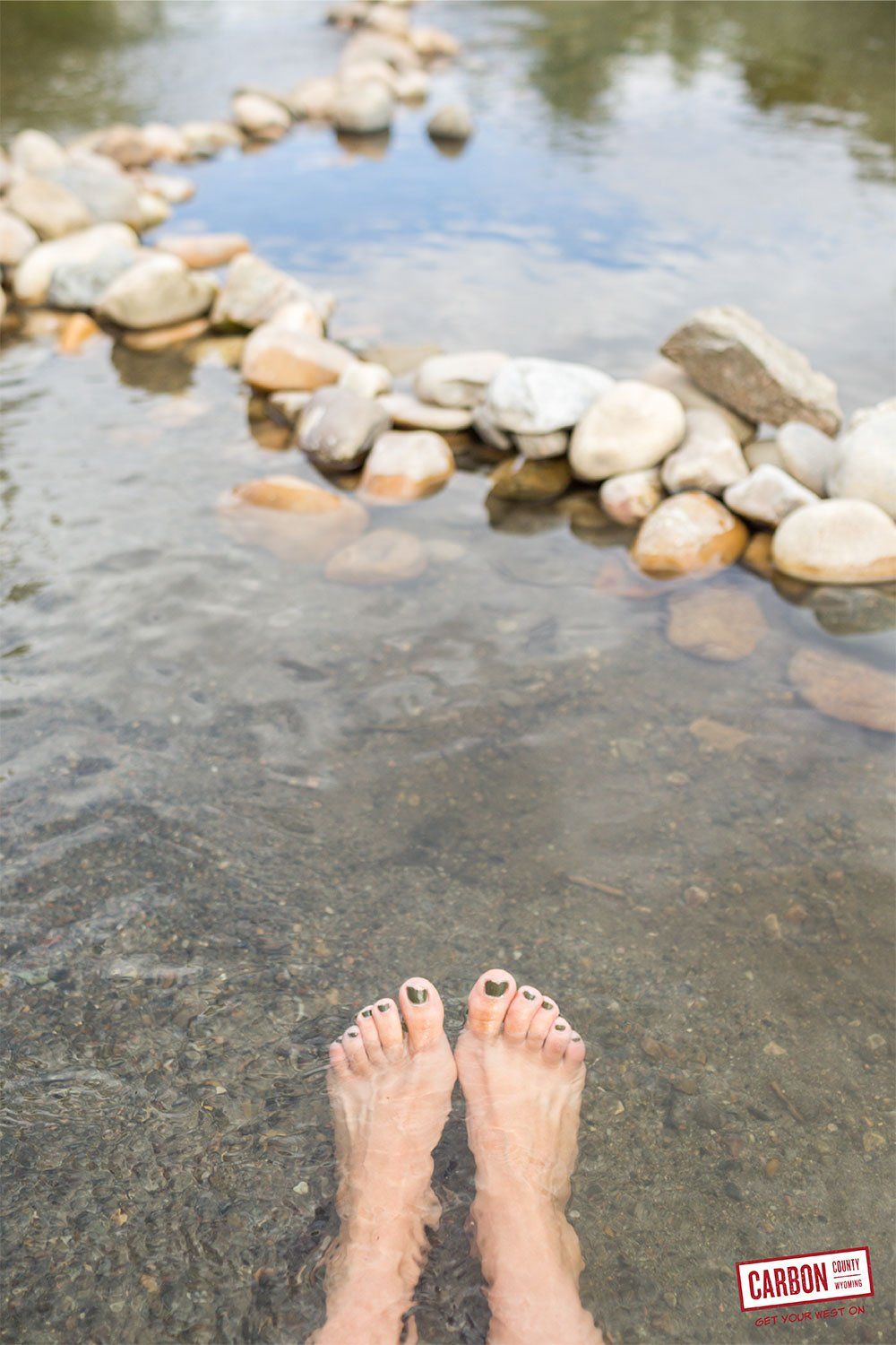 A person 's feet are standing in a body of water surrounded by rocks.